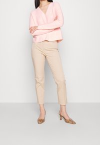 Pull léger rose, à col en V, avec des manches longues ; assorti à un pantalon beige, ajusté, et des chaussures à talons hauts beiges et texturées.
