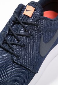 Dunkelblaues Textil-Sneaker mit strukturierter Oberfläche, schwarzen Schnürsenkeln, einem orangefarbenen Logo-Patch und einer weißen Gummisohle.