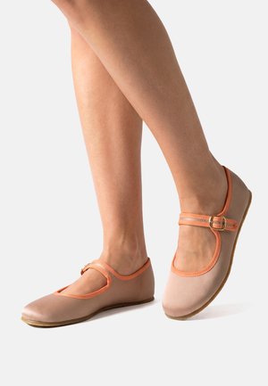 Gambe con scarpe Mary Jane beige con rifiniture arancioni e fibbie su uno sfondo chiaro uniforme.