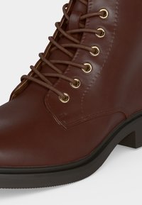 Bottine en cuir marron à bout rond, œillets dorés, lacets marron et talon et semelle en caoutchouc noir bas.