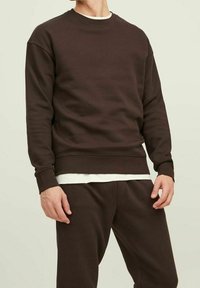 Brun bomullströja med rund halsringning, ribbade ärmslut och nederkant, bärs över en vit skjorta, del av matchande byxor.