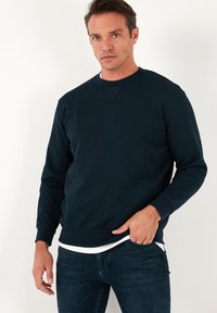 Tengerészkék crewneck pulóver, pamut keverékből készült, hosszú ujjaival, bordázott mandzsettákkal és finom varrási részlettel a nyakkivágásnál.