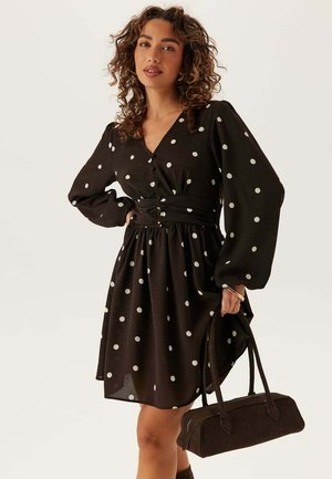 Kvinde med krøllet hår iført sort kjole med prikker, der holder en sort håndtaske mod en ensfarvet baggrund.