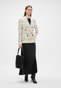 Γυναίκα με ίσια καστανά μαλλιά, ντυμένη με λευκό σακάκι διπλής όψεως με μαύρο σχέδιο, μακριά μαύρη φούστα, μαύρες μπότες με τακούνι, κρατώντας μαύρη τσάντα.