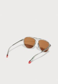Ochelari de soare cu nuanță maronie și ramă transparentă. Brațele au vârfuri roșii, accente metalice și o formă rotundă. Obiectivul oferă o estetică vintage.