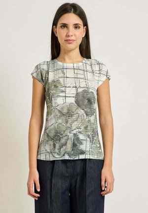 Donna che indossa una maglietta beige a maniche corte con stampa floreale verde e linee a griglia nere, abbinata a pantaloni blu scuro, in piedi davanti a uno sfondo semplice.