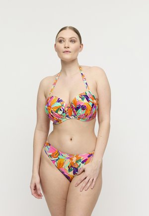 Femme portant un bikini à fleurs coloré avec un col en halter, debout avec une main reposant sur sa cuisse, regardant légèrement sur le côté.