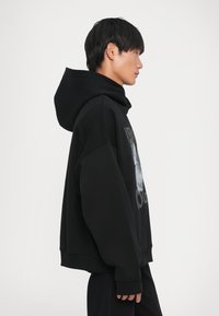 Melnā kapuce džemperis ar lielu kapuci, garām piedurknēm un grafisku druku priekšā. Mīksta tekstūra un pārāk liela piegriezuma.