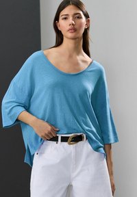 Top azzurro chiaro oversize realizzato in tessuto morbido, con scollatura ampia e maniche corte, abbinato a pantaloni bianchi a vita alta e una cintura nera.