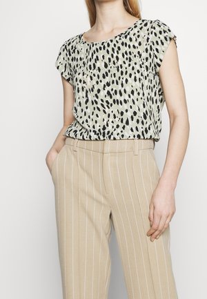 Vrouw draagt een blouse met korte mouwen en dierenprint, ingestopt in beige broek met fijne strepen, met één hand in de zak.