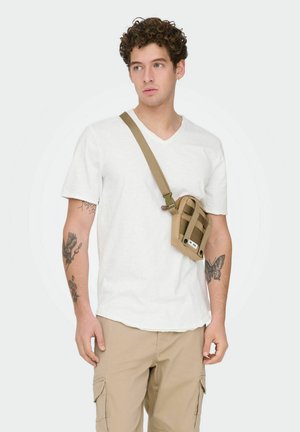 Young man with tattoos wearing white t-shirt, beige cargo pants, and a tan crossbody bag, looking slightly to his left.