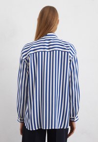 Chemise à rayures avec des rayures verticales bleues et blanches, un col classique et des manches longues avec des poignets. Tissu lisse, coupe décontractée.
