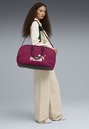 Dufflebag in levendig magenta met zwarte accenten, voorzien van bovenhandvaten en een schouderriem. Groot "PUMA" logo en een elegante, afgeronde vorm.
