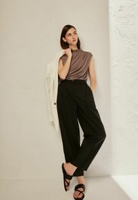 Hnědý draped top s krátkými rukávy, černé kalhoty s širokými nohavicemi a černé sandály; doplněno bílým blejzrem přehozeným přes jedno rameno.
