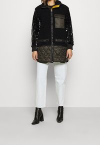 Svart fleece- och vadderad jacka med blanka svarta ärmar, dragkedjefickor och en gul krage. Bärs över ljusa jeans och stövlar.