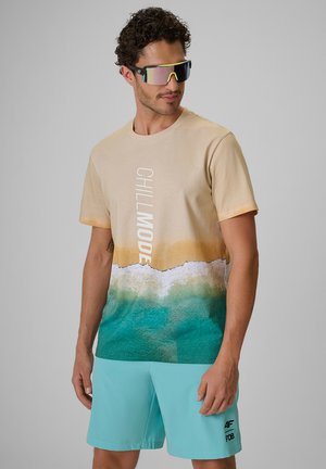 Mężczyzna noszący odblaskowe sportowe okulary przeciwsłoneczne, beżowy t-shirt z motywem oceanu "CHILL MODE" i jasnoblękitne spodenki na szarym tle.