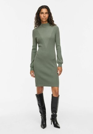 Femme aux cheveux bouclés portant une robe en maille verte à manches longues et des bottes noires à talons montant jusqu'aux genoux, debout devant un fond blanc uni.