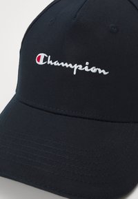 Fekete sapka pamut keverék anyagból, fehér hímzett "Champion" logóval és piros díszítéssel a bal oldalon. Strukturált kialakítás, ívelt karimával.