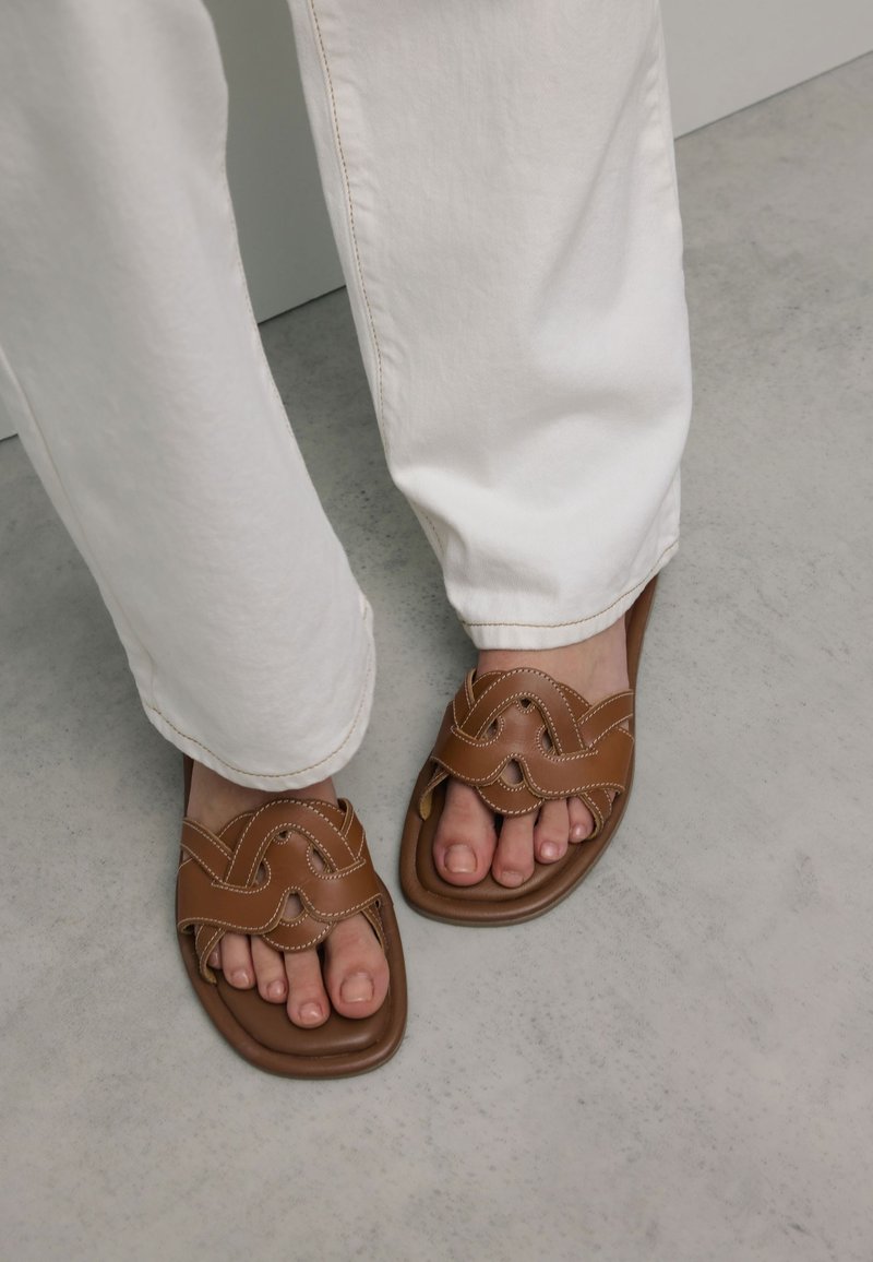 Sandales à glissière en cuir marron avec un design de sangle ondulée et des coutures blanches contrastées, assorties à un pantalon large blanc sur un sol gris.