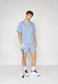Chemise à manches courtes et short assorti en bleu avec des motifs géométriques circulaires. Gants de golf et chaussures blanches, avec un club de golf.