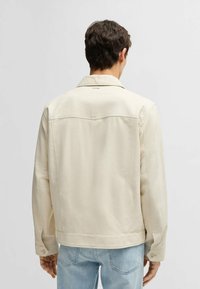 Beige bomullskavaj med en strukturerad krage, knäppta manschetter och en ensam synlig logotyp på övre ryggen. Har en slät textur.