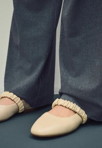 Beige nahkakengät, joissa on terävä kärki ja rypytetty nilkkareuna, yhdistettyinä löysiin tummanharmaisiin housuihin. Teksturoitu pinta näkyvissä.