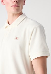 Kremasta polo majica s texturetiranom tkaninom, klasičnom kragnom i malim CK logotipom na lijevoj strani prsnog koša. Kratki rukavi, zatvaranje na dugmad.