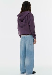 Felpa con cappuccio viola con stampa grafica sul retro, abbinata a jeans di denim azzurri a vestibilità morbida. Tessuto liscio, silhouette rilassata.