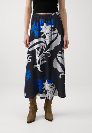 Femme portant une jupe midi bleu marine avec de grandes impressions florales blanches et bleues, associée à des bottes beiges à bout pointu et un haut noir sans manches.