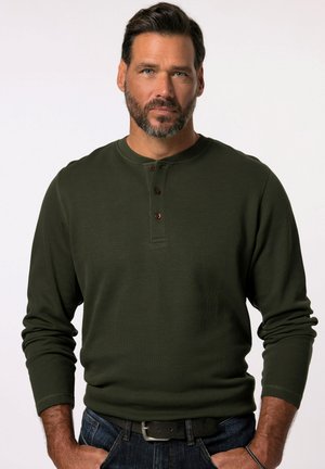 Camisa henley de manga larga de color verde oscuro con cuello redondo, textura acanalada y tres botones marrones en el pecho. Se lleva con unos vaqueros azules.