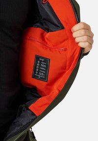 Zielona wodoodporna kurtka z pomarańczową podszewką. Posiada wewnętrzną kieszeń na suwak oraz metkę pielęgnacyjną przyszytą do podszewki.