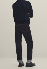 Tummansiniset farkut suoran leikkauksen kanssa, joissa on käännetyt lahkeet, kontrastiompeleet ja klassiset takataskut, yhdistettynä yksinkertaiseen tummansiniseen takkiin.