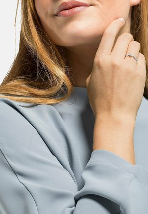 Silberner Ring mit einem kleinen Edelstein, getragen an einer Hand mit mittlerem Hautton. Grauer Stoffärmel teilweise sichtbar, langes, glattes Haar berührt die Schulter.