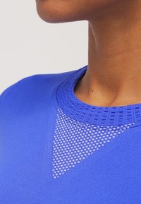 Camiseta deportiva azul con escote redondeado texturizado y panel de malla con puntos blancos. Tejido suave y diseño ajustado cerca del cuello.