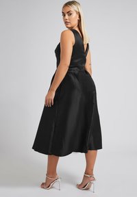 Mustat satiini mekko, jossa on hyvin istuva korsetti, v-mallinen selkäosa ja levenevä polvipituinen hame. Siinä on sivutaskut ja sileä pinta. Korkokengät näkyvissä.