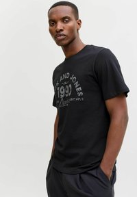 Juodas medvilninis marškinėliai su trumpomis rankovėmis, su spausdintu tekstu nusidėvėjusių stiliumi. Modelis pozuoja su rankomis kišenėse, vilkėdamas tamsias kelnes.