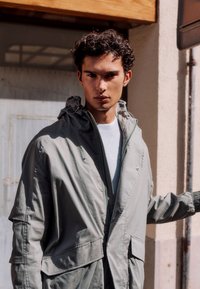 Mladý muž s kudrnatými vlasy, který má na sobě světlou šedou bundu s kapucí a bílé tričko, stojí venku u vchodu do budovy.