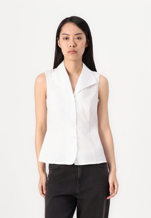 Blouse sans manches blanche avec un col en V, boutons à l'avant, design ajusté et texture lisse, associée à un jean foncé.