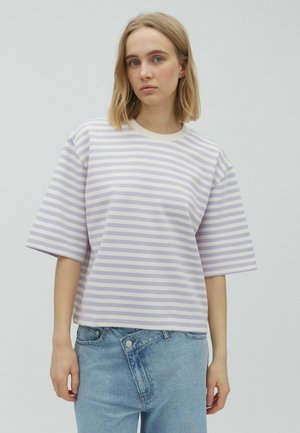Ung kvinde med blondt hår iført en kortærmet lavendel- og hvidstribet skjorte og asymmetriske blå jeans foran en ensfarvet baggrund.