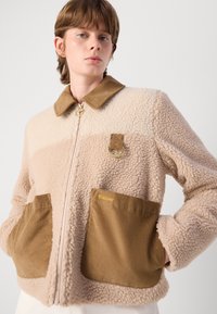 Veste en polaire beige avec un haut crème. Dotée d'un col en velours côtelé et de poches, de quincaillerie dorée et d'un logo doré sur la poche gauche.