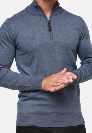 Homme portant un pull bleu à fermeture éclair quart, mains jointes devant, debout sur un fond uni.