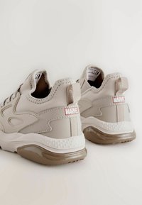 Baskets beiges avec tige synthétique lisse, col rembourré et semelle en caoutchouc marron. Présente un logo rouge "MARVEL" sur l'onglet du talon.