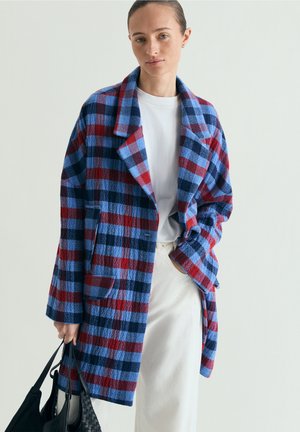 Mujer que lleva un abrigo a cuadros azul, rojo y negro sobre una camisa y falda blancas, sosteniendo un bolso negro sobre un fondo liso.