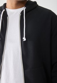 Schwarze Zip-Hoodie mit weißen Kordeln, aus weichem Baumwollmaterial und einer Fronttasche. Getragen über einem schlichten weißen T-Shirt.