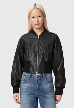 Młoda kobieta o długich blond włosach nosi czarną skórzaną kurtkę typu bomber i niebieskie dżinsy, stoi na tle jednolitego szarego tła.