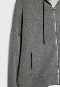 Sweat à capuche gris zippé avec poche avant, capuche à cordon réglable et détail de fermeture éclair argentée visible.