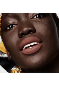 Gros plan sur un visage à la peau foncée, avec des lèvres nues mates et des dents blanches. Un foulard à motifs est partiellement visible en arrière-plan.