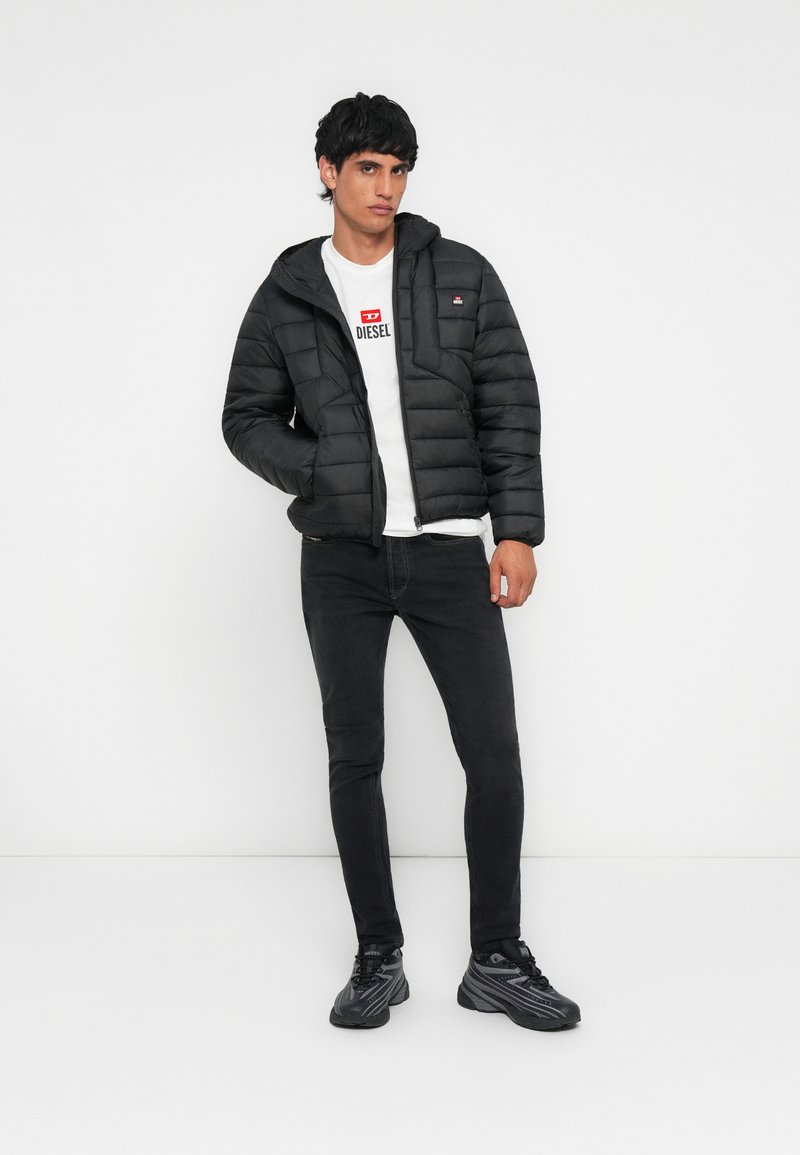 Man in een zwarte pufferjack, een wit t-shirt met het Diesel-logo, zwarte skinny jeans en zwarte sneakers, staand tegen een witte achtergrond.