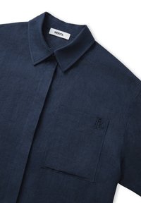 Marineblaue Leinenbluse mit klassischem Kragen, ausgestattet mit einer Brusttasche und gesticktem Logodetail. Glatte Textur und lässige Passform.