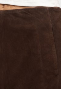 Gros plan sur une jupe en daim marron foncé portée avec un haut blanc, mettant en avant la ceinture et la texture du tissu.